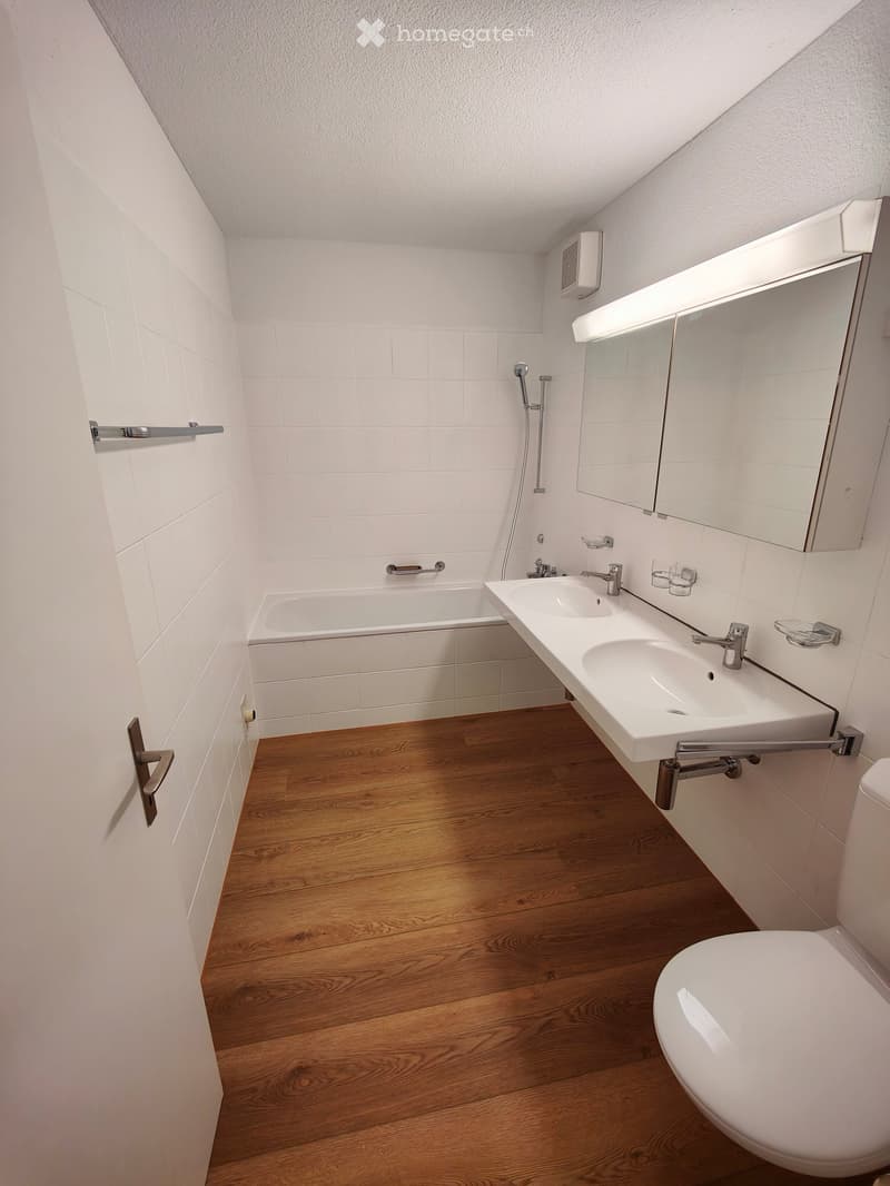 Modern bathroom with white tiles, wooden floor, bathtub, double sink, toilet, and large mirror with built-in lighting.