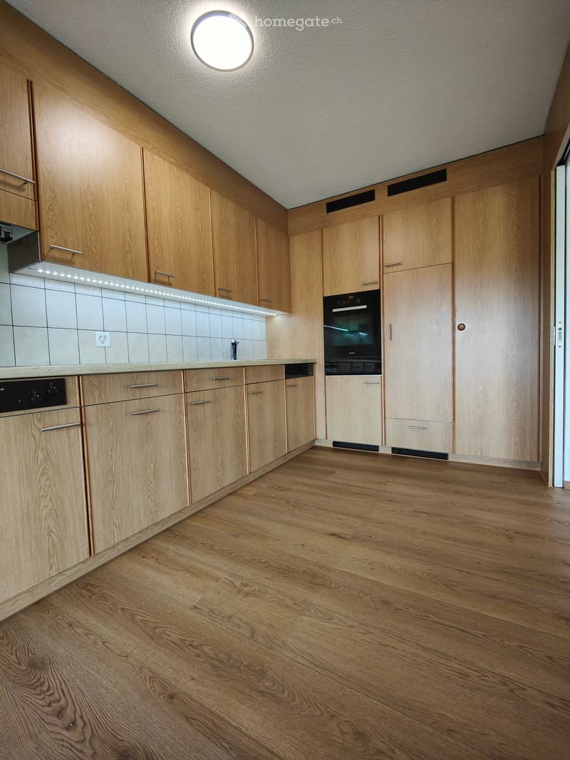 Modern kitchen with wooden cabinets, light wood flooring, and built-in appliances including an oven and dishwasher.