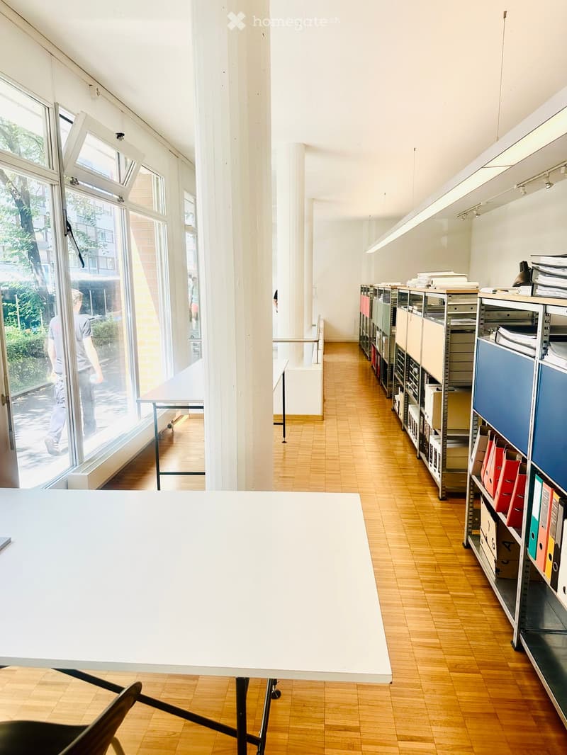 Bright, open room with wooden flooring, large windows, a white table, a chair, and shelves filled with files and binders.