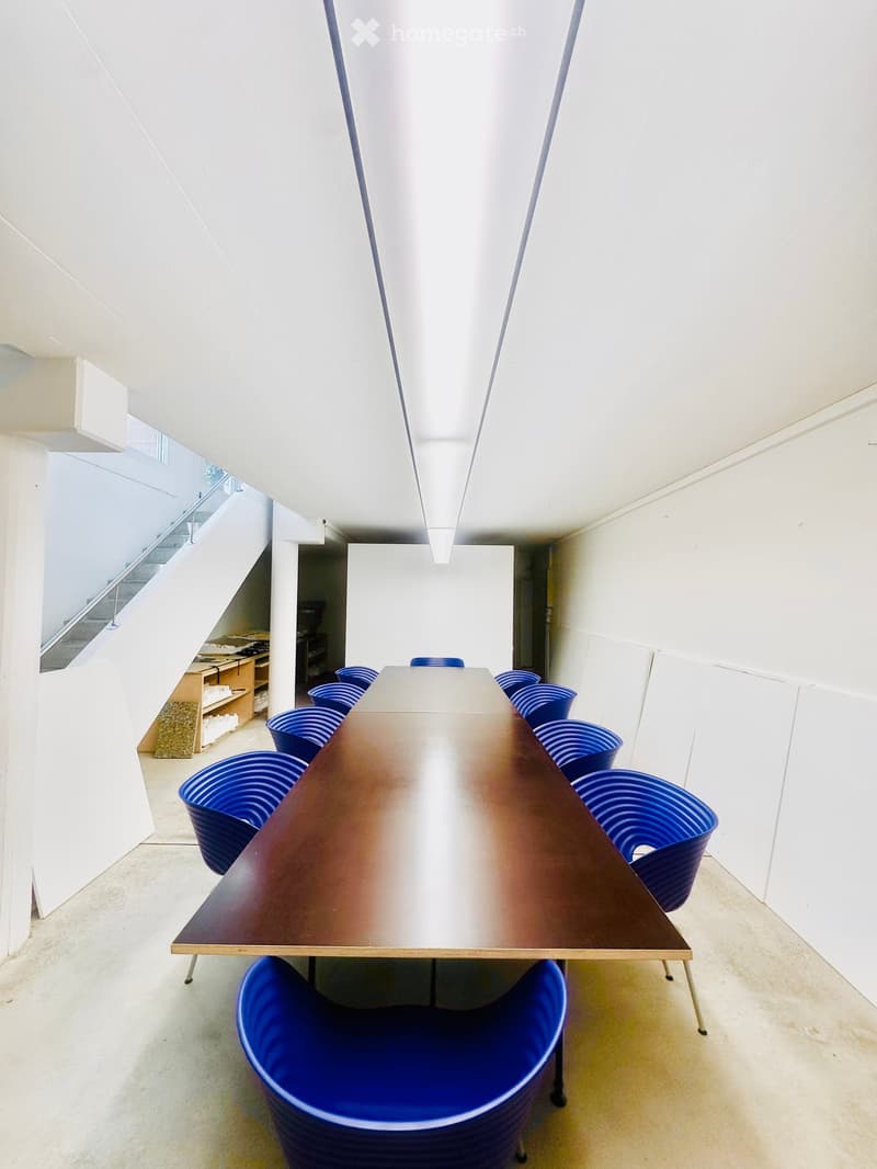A modern, minimalist room with a long wooden table and several blue chairs arranged around it. The space features white walls, a clean and open layout, and a visible staircase in the background.