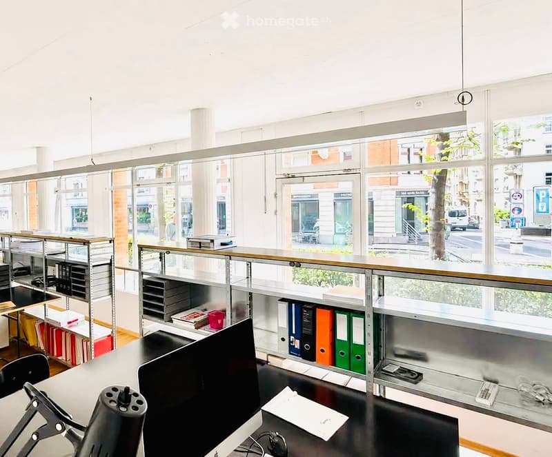 Modern office space with large windows, a desk with a computer screen, office chair, shelves with books and folders, and a glass partition.