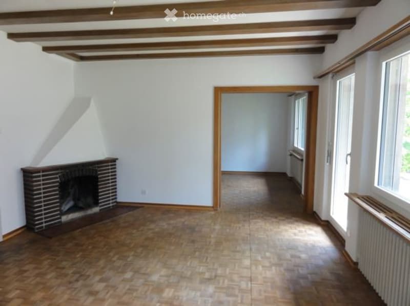 Empty room with wooden flooring, white walls, exposed wooden ceiling beams, and a brick fireplace. There is a doorway leading to another room.