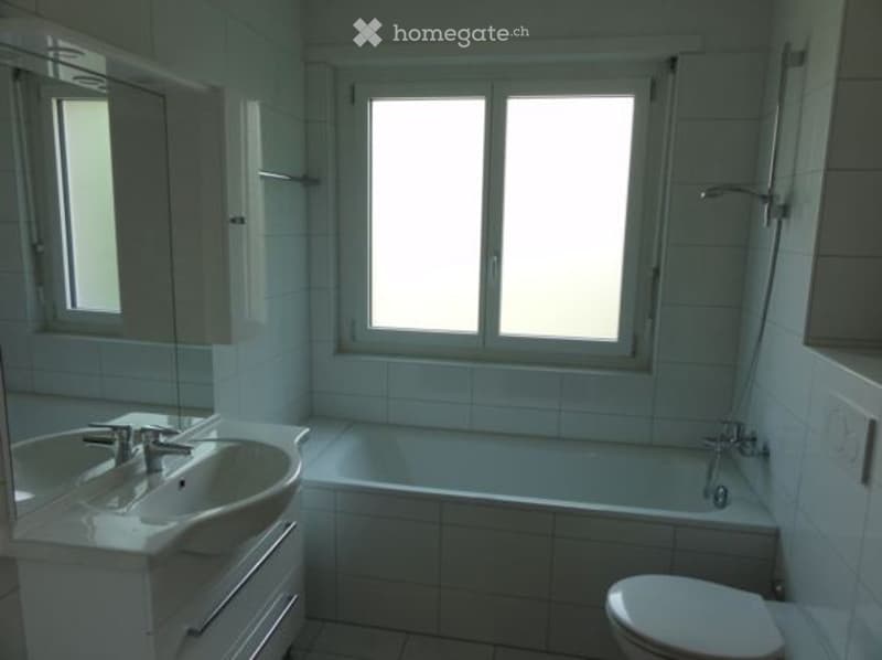 Modern bathroom with white tiles, a sink with a faucet, a bathtub, and a toilet. The room has large windows providing natural light.