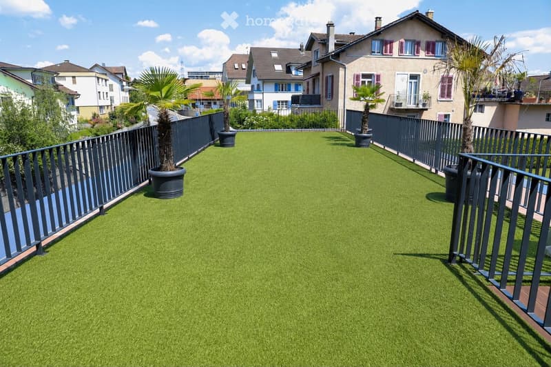 A rooftop garden with artificial grass, surrounded by a metal railing and potted plants, with residential buildings in the background.