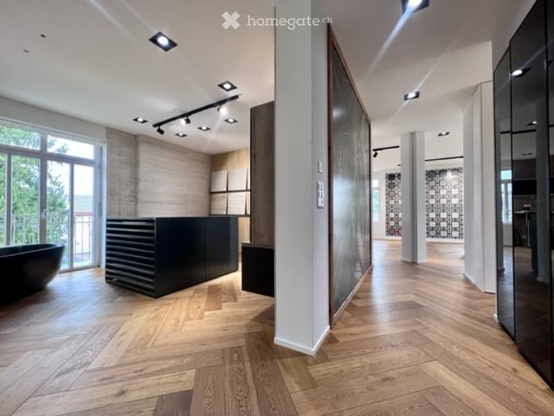 Modern, minimalist room with wooden herringbone flooring, large windows, and a black reception desk. A bathtub is visible in the foreground, and the space features recessed lighting and a balcony view.