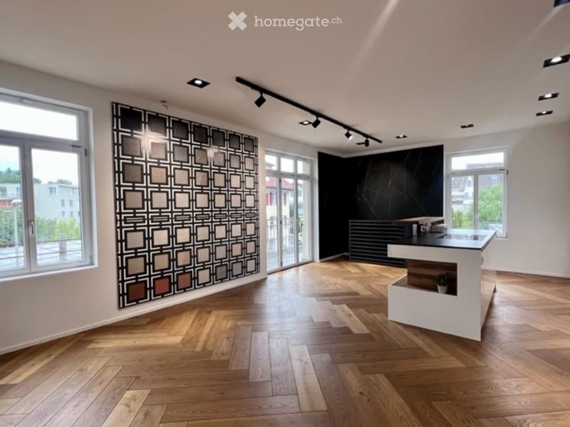 Modern reception area with herringbone wooden flooring, white reception desk, decorative wall art, and large windows offering a view of the outside.