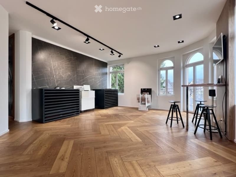 Modern, minimalist room with a reception desk, two stools, a table, and a screen. The room features large arched windows, wooden flooring, and track lighting.
