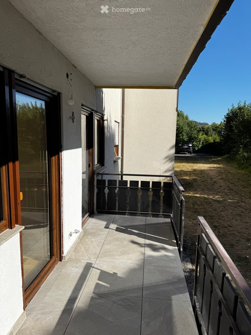 Balcony with tiled floor, wooden-framed glass door, black metal railing, and view of greenery and a parked car.