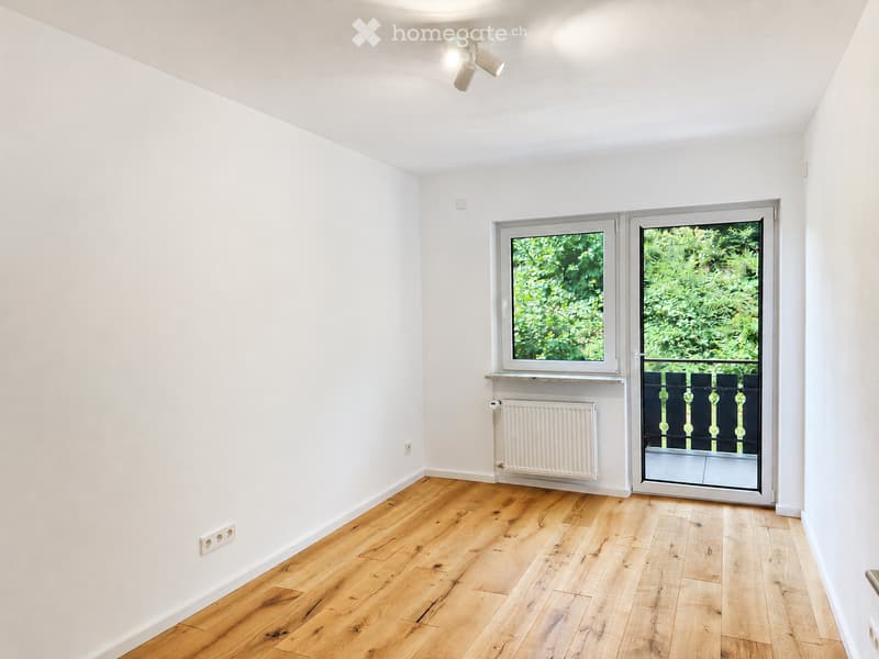 Empty room with light wooden flooring, white walls, and a large window with a view of greenery. A radiator is visible below the window, and a sliding glass door leads to a balcony area.