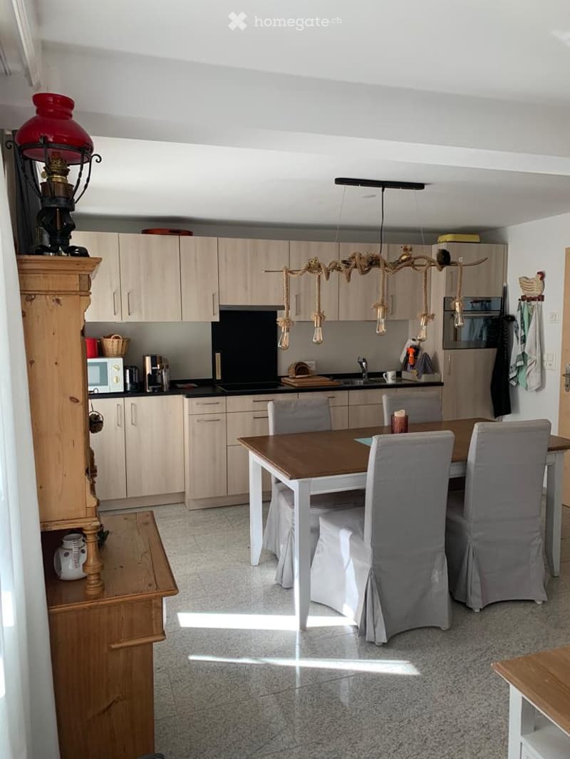 Bright kitchen with light wood cabinets, a central dining table with chairs, a microwave, and a hanging light fixture.