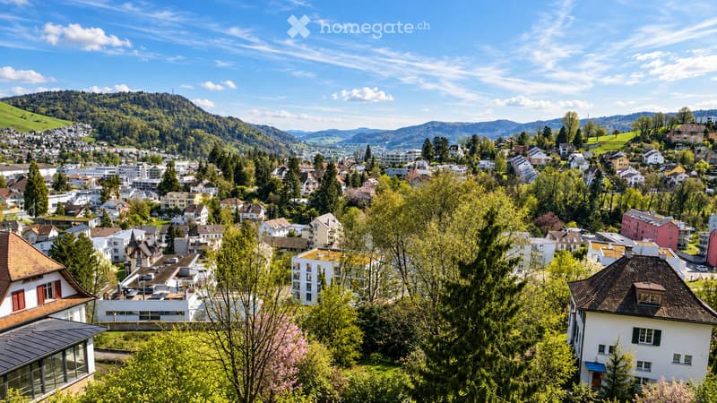 A panoramic view of a town nestled in a valley with a mix of residential and commercial buildings, surrounded by lush green hills and a clear blue sky.