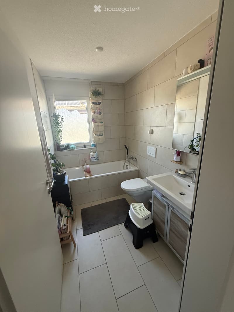 Modern bathroom with a bathtub, toilet, sink, and a small stool. Features tiled walls and floor, a window with a plant, and a mirror above the sink.