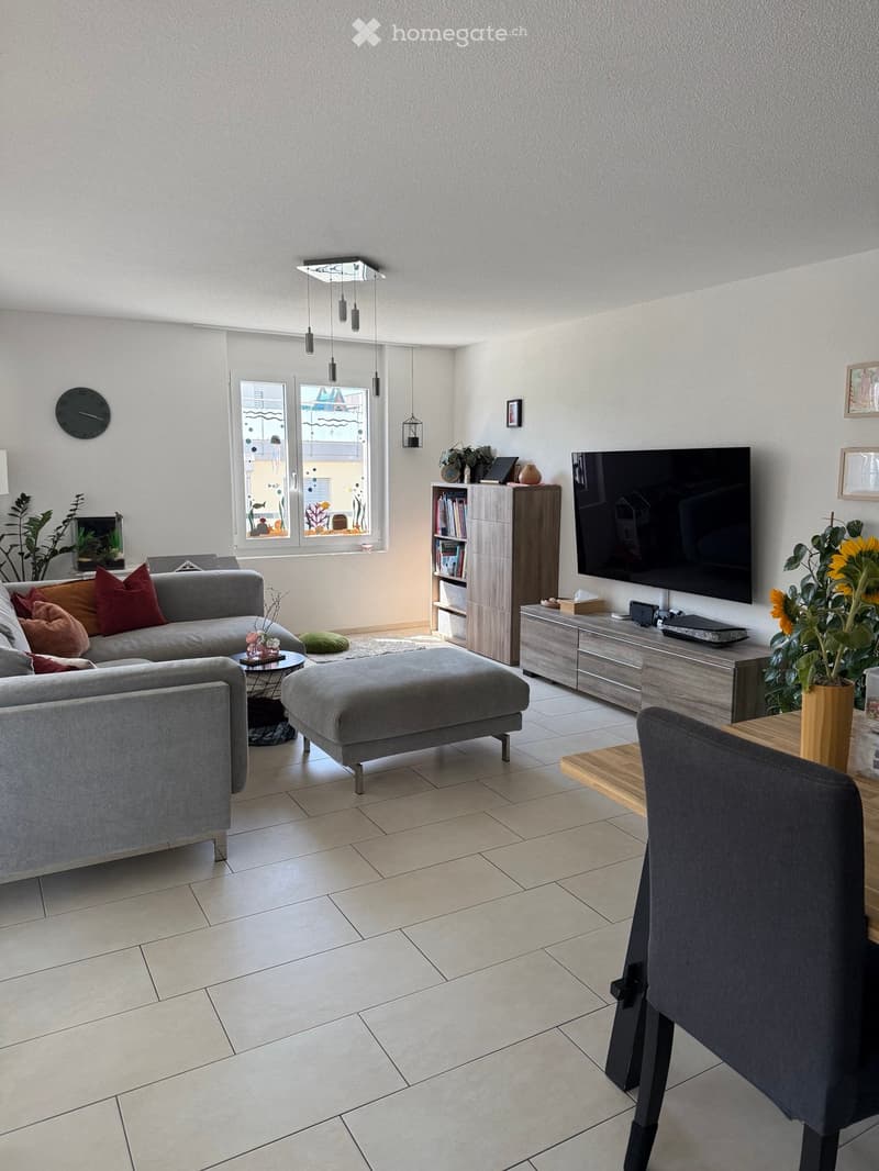 Modern living room with grey sofa, ottoman, coffee table, and a television mounted on the wall. The room features a bookshelf, a small table with a plant, and a large window with a view.