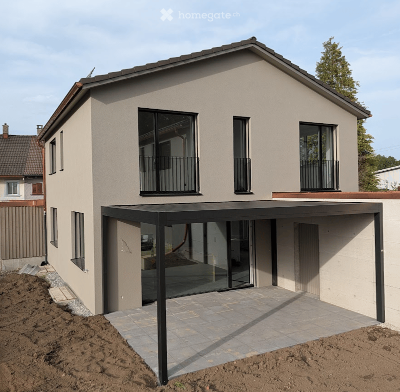 Modern two-story house with beige facade, large windows, and balconies. Features a covered patio area with glass walls and a tiled floor.
