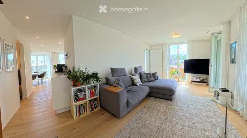 Modern living room with grey sectional sofa, coffee table, bookshelf, dining area, and large windows with a view.