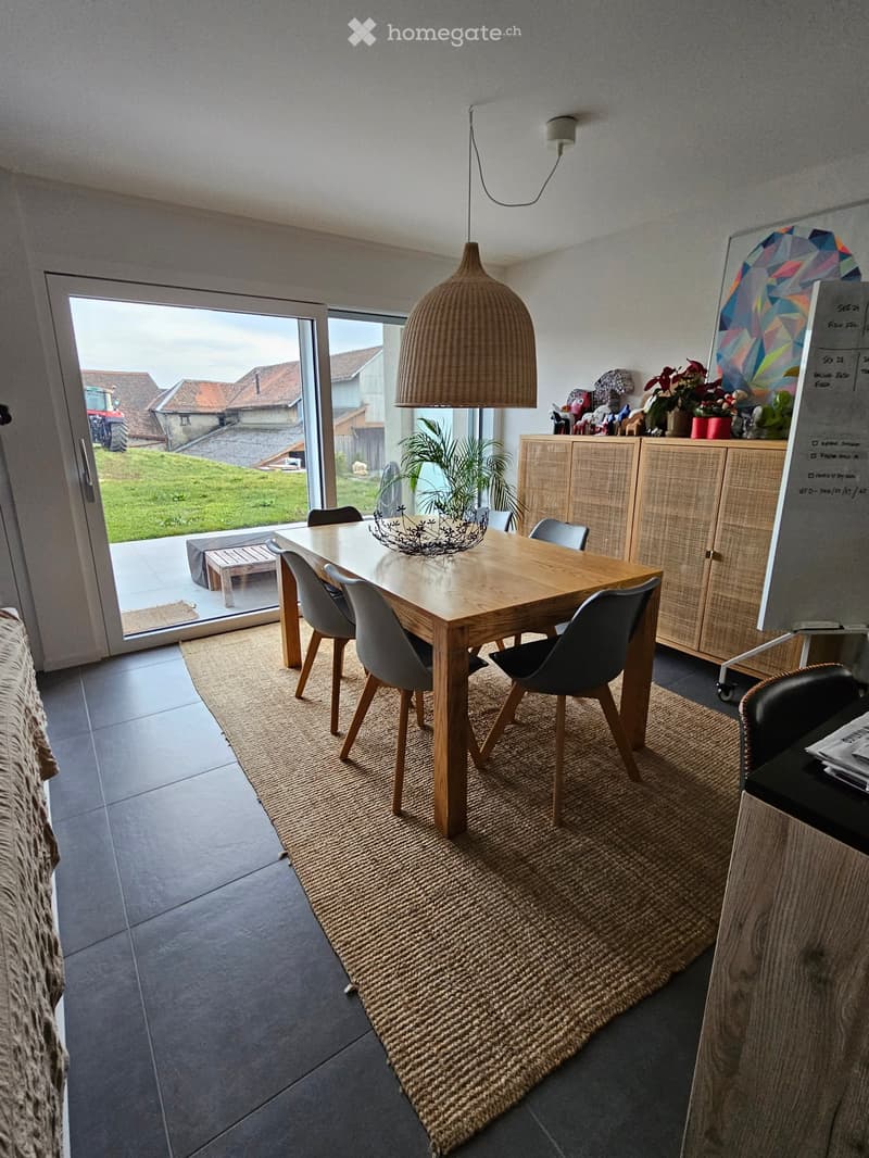 Dining room with a wooden dining table, six chairs, a pendant light, a cabinet, and a plant on the table. The room has large windows with a view of a green lawn and a tractor outside.