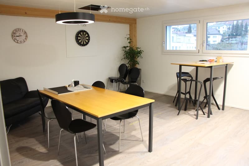 Bright room with a sofa, two tables with chairs, a dartboard, a clock, and a plant. Large windows provide natural light.