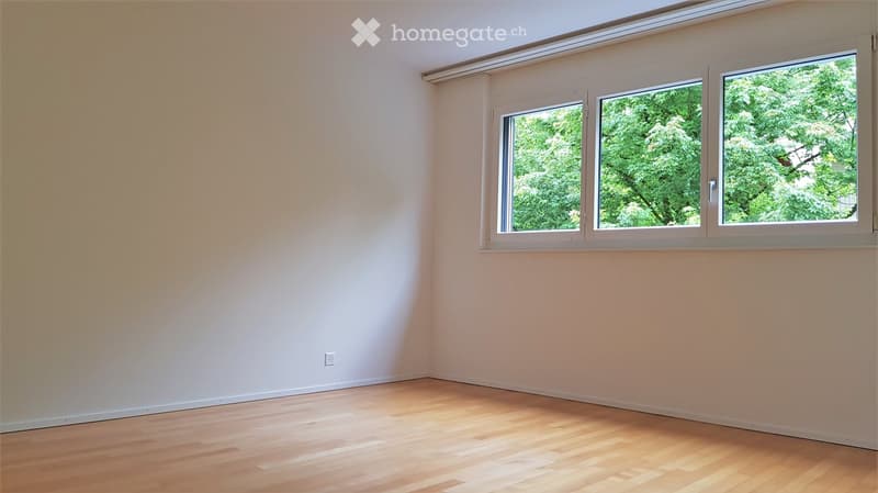 Empty room with light-colored walls and wooden flooring, featuring large windows with white frames and a view of greenery outside.