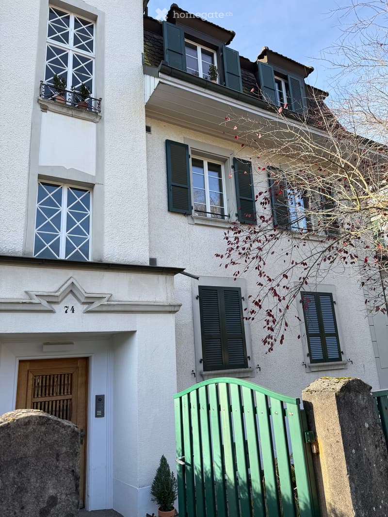 Two-story house, white exterior, black shutters, balcony with plants, wooden door, green gate
