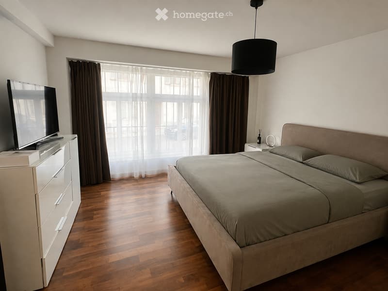 Modern bedroom with a large bed, nightstand, television, and large windows with curtains. The room features wooden flooring and a hanging pendant light.