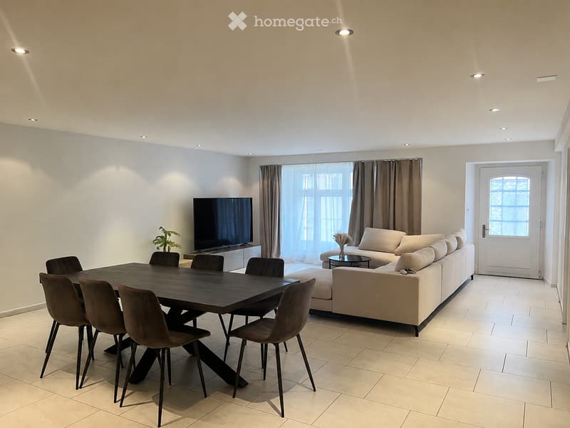 Modern living and dining area with a large sectional sofa, coffee table, flat-screen TV, dining table with six chairs, and a small side table with a vase.