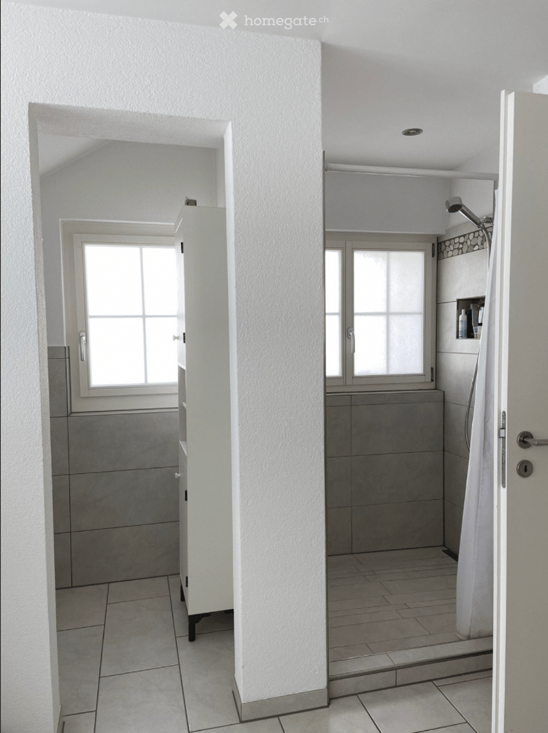 Modern bathroom with tiled walls and floor, shower area with a curtain, small window, and a cabinet with a bottle on top.