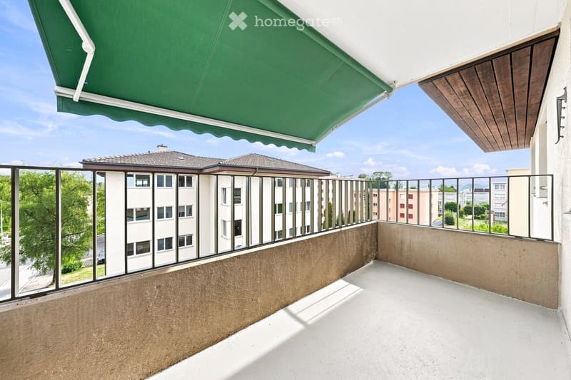 Balcony with a green awning, metal railing, and a view of a residential area with buildings and trees.