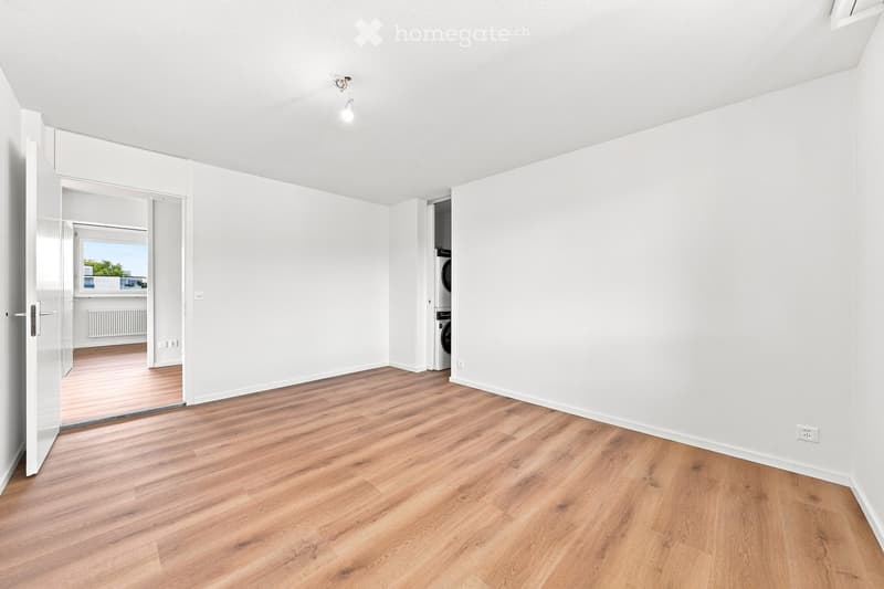 Empty room with light wooden flooring, white walls, and a single ceiling light fixture. An open doorway leads to a balcony area with a view.