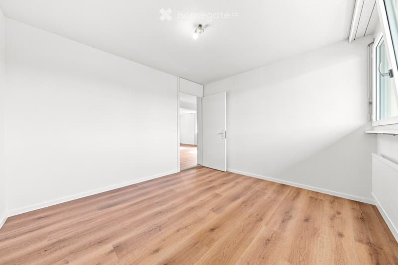 Empty room with white walls, wooden flooring, and a door leading to another space.