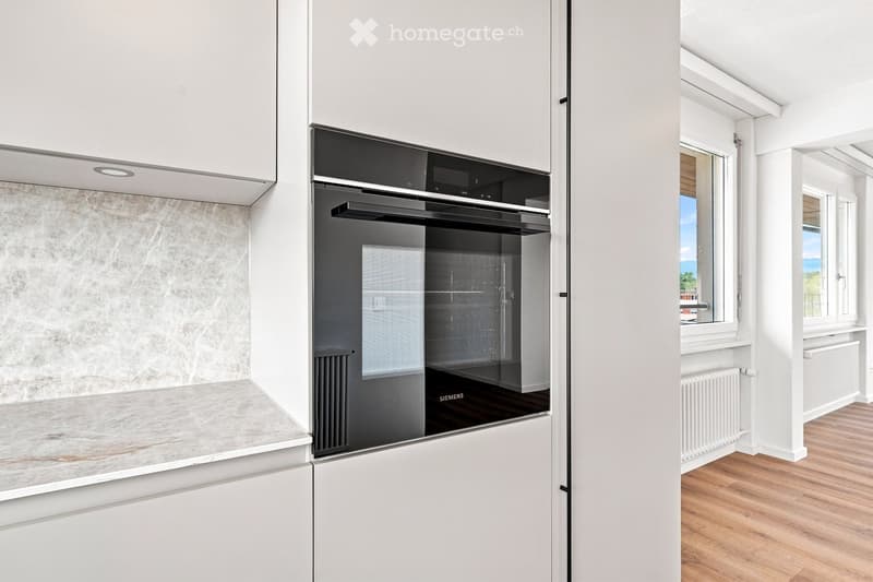 Modern kitchen with white cabinets, a built-in Siemens oven, and a marble backsplash. The space features wooden flooring and a window offering a scenic view.