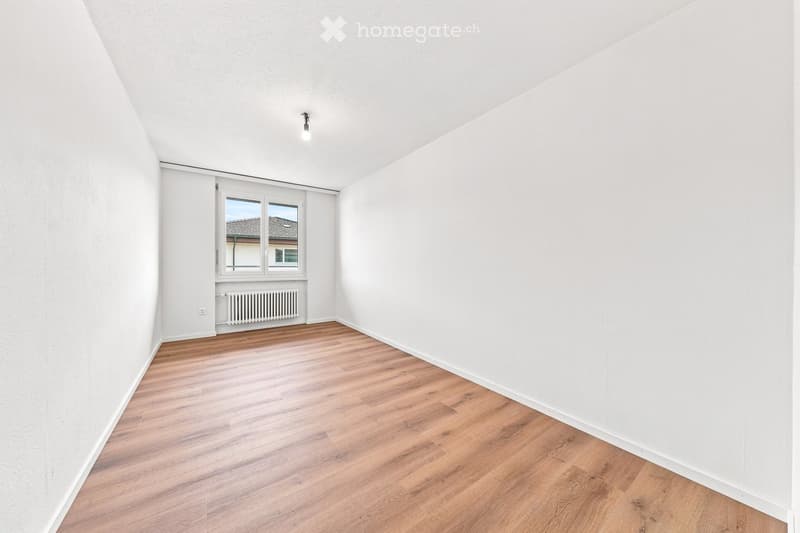 Empty room with white walls, wooden flooring, and a window with a radiator below it.
