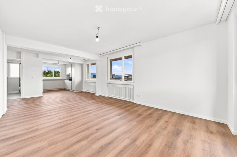 Bright, unfurnished room with wooden flooring, white walls, large windows, and a view of the outdoors. The room is connected to a small kitchen area with white cabinets and a countertop.