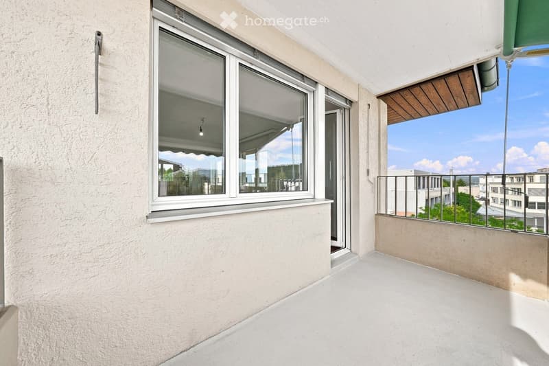 Balcony with white concrete floor, metal railing, large window with white frame, and wooden awning.