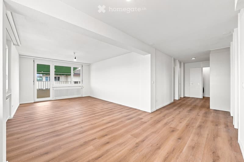 Empty room with wooden flooring, white walls, and large windows leading to a balcony.