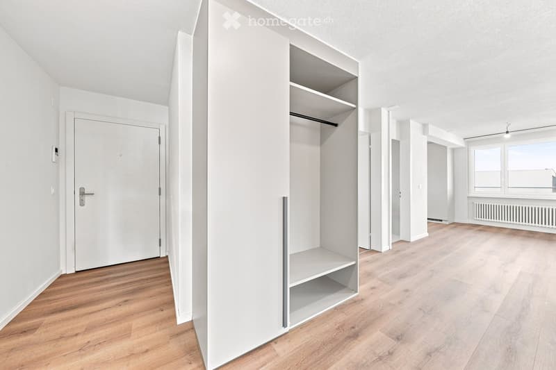 Empty room with light wooden flooring, white walls, built-in shelving unit, and large windows allowing natural light.