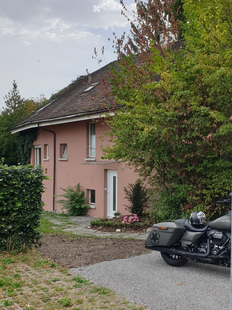 Single-story pink house with white-framed windows and a white front door, surrounded by greenery and a gravel driveway with a parked motorcycle.