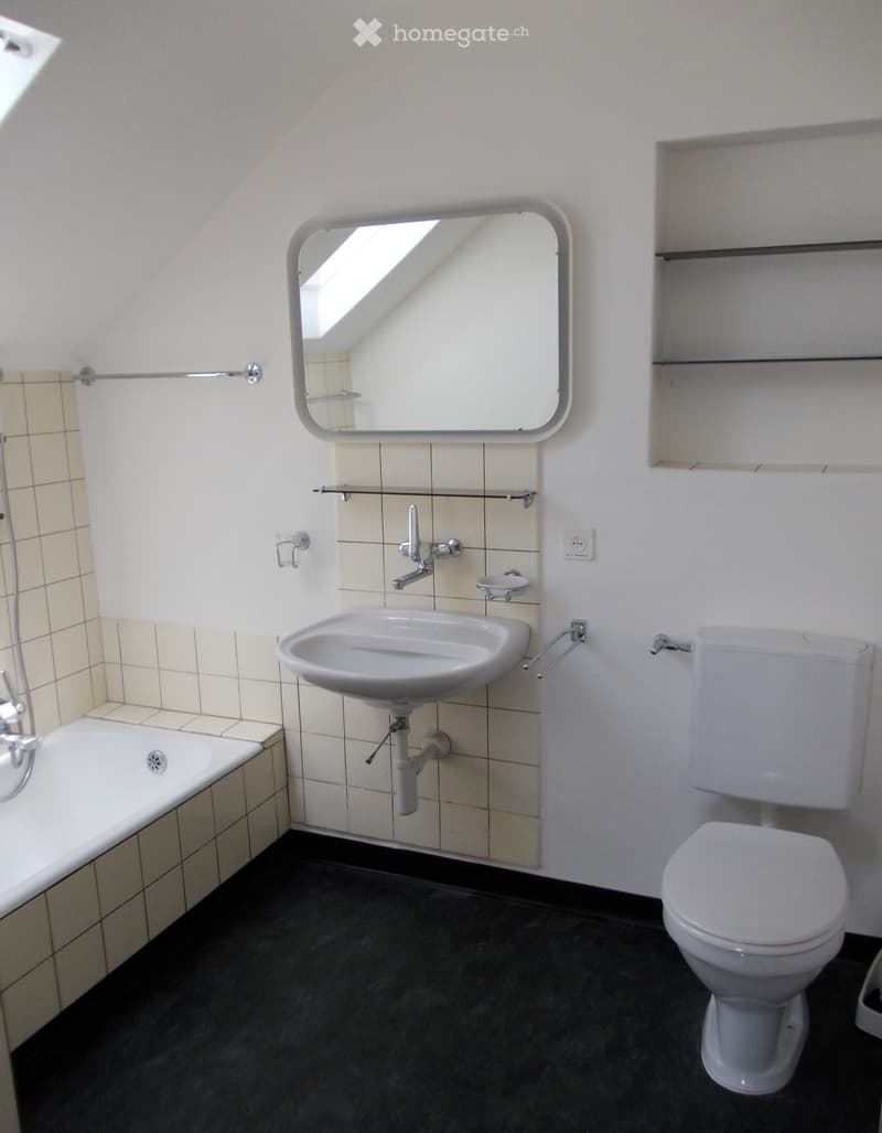 Modern bathroom with a white sink, toilet, bathtub, and large mirror above the sink. The walls are tiled, and the floor is dark.