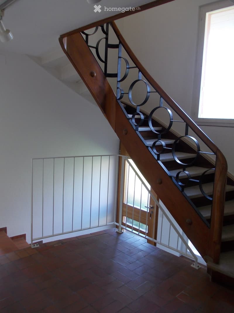 Staircase with wooden steps and metal railing, leading to an open doorway with glass panels. The floor is tiled with reddish-brown tiles.