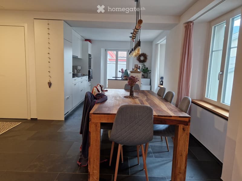 Bright, modern dining area with a long wooden dining table surrounded by grey upholstered chairs. The space features a sleek kitchen area with white cabinets, an oven, and a faucet. Natural light streams in through large windows with pink curtains, and de