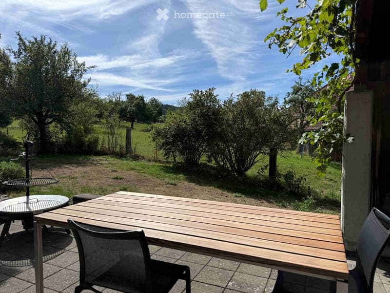 Outdoor patio with wooden table, chairs, and a view of a green landscape with trees and hills.