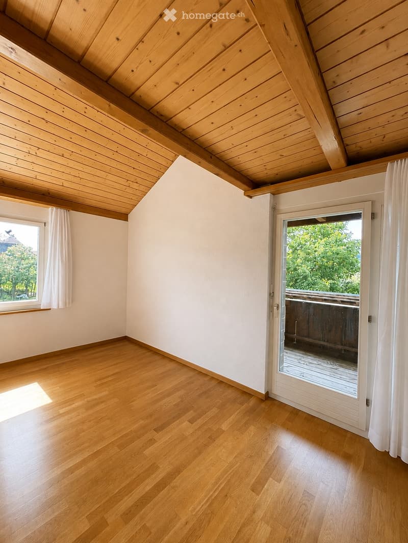 Empty room with wooden flooring, white walls, and a slanted wooden ceiling. Features a large window with white curtains and a sliding glass door leading to a balcony.