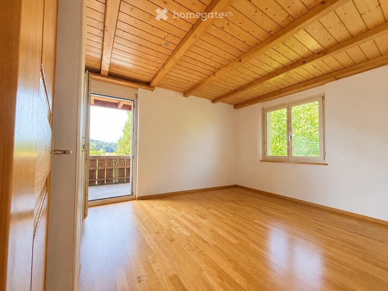 Empty room with wooden flooring, white walls, exposed wooden ceiling beams, large window, and sliding glass door leading to a balcony.