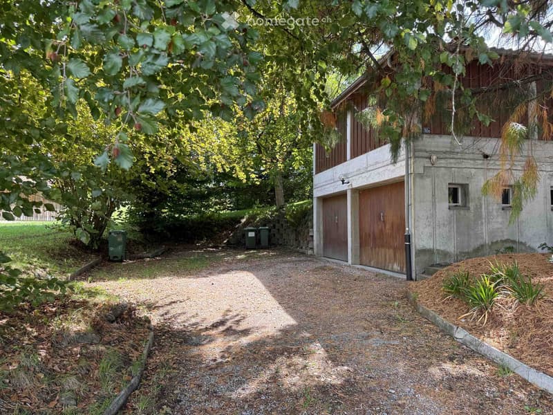 A small, two-story building with a wooden garage door and concrete walls, surrounded by a gravel pathway and greenery.
