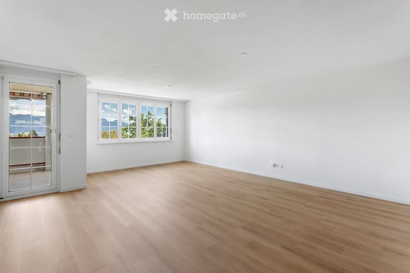 Empty room with light wooden flooring, white walls, large windows, and a sliding glass door leading to a balcony.