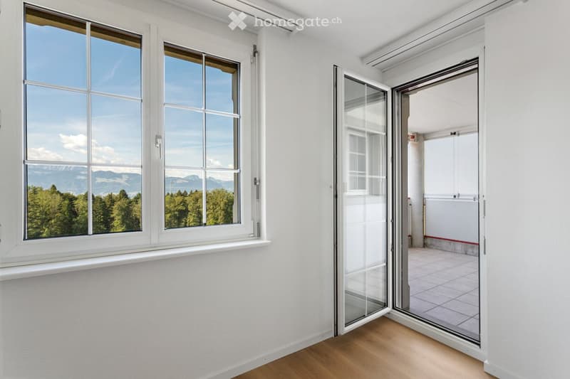 Empty room with white walls, wooden flooring, large windows, and an open sliding glass door leading to a balcony or terrace.