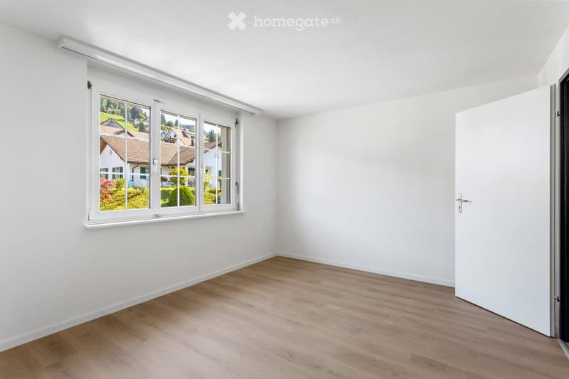 Empty room with white walls, wooden flooring, and a large window showing a view of houses and greenery outside.