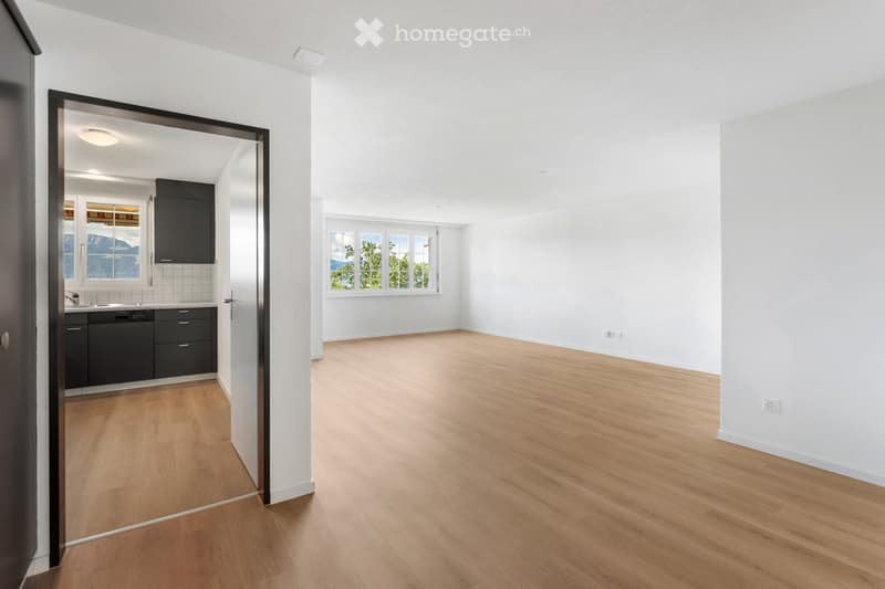 Empty room with light wooden flooring and white walls, featuring large windows and a view of greenery. A partially visible kitchen area with cabinets and a sink is seen through an open doorway.