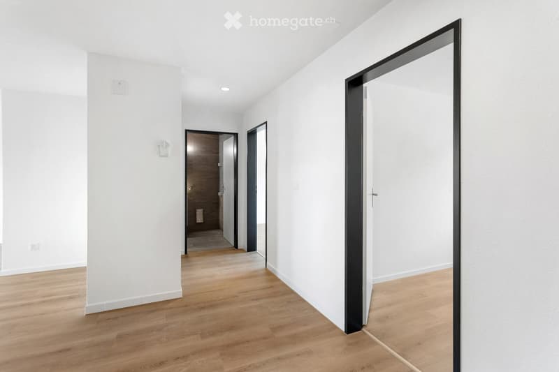 Bright, minimalist hallway with light wooden flooring, white walls, and recessed ceiling lighting. Features a large black-framed mirror on the right and a wooden door at the end.
