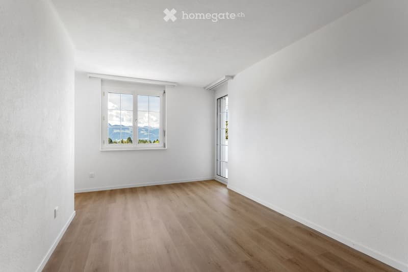 Empty room with white walls, wooden floor, large window with scenic view, and glass door leading outside.