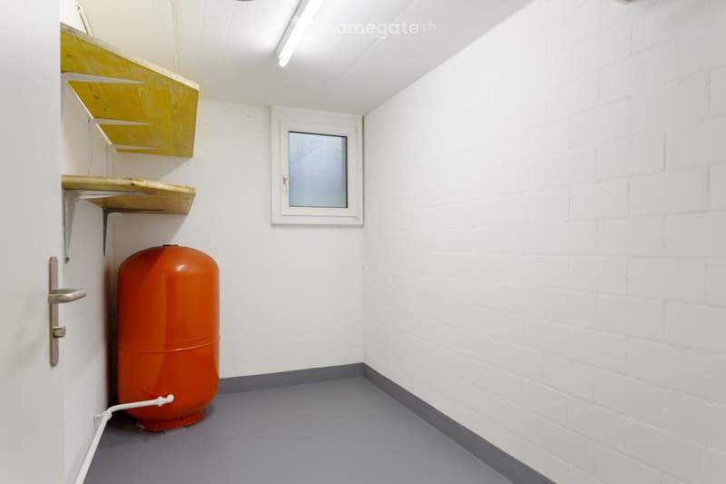 Completely empty room with white brick walls, grey floor, and a small window. A large orange cylindrical object is present, likely a water tank or boiler, but no furniture or equipment.
